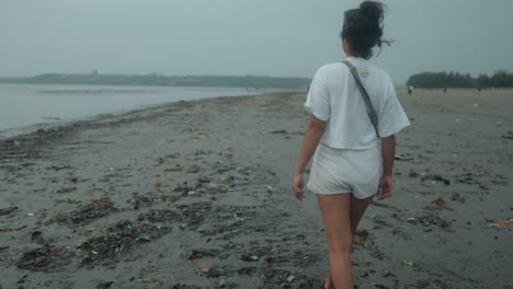 Medium-slow-motion-dolly-shot-of-a-young-indian-woman-in-white-shirt-and-fanny-pack-while-taking-an-evening-walk-on-the-beach-during-her-summer-vacation-with-a-view-on-beautiful-beach-with-calm-sea