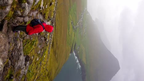 Woman-in-red-raincoat-looks-over-foggy,-dramatic-Faroe-Islands-landscape
