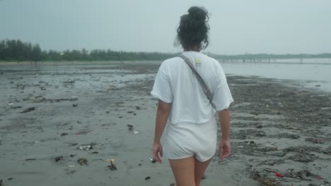 Medium-dolly-shot-of-a-young-indian-woman-in-white-shirt-and-fanny-pack-while-taking-an-evening-walk-on-the-beach-during-her-summer-vacation-in-slow-motion