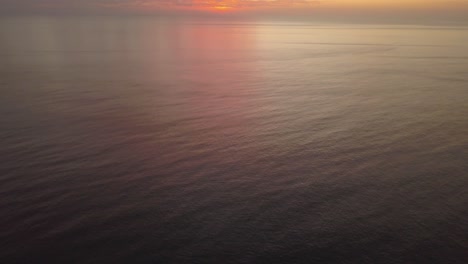 Hypnotic-black-water-of-the-ocean-during-sunset,-magical-pink-sky-pan-up-over-the-horizon-drone-shot