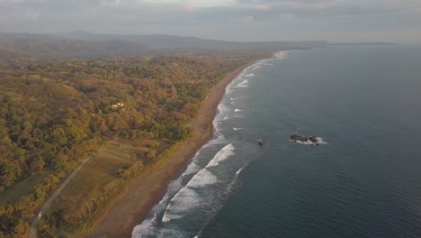 Majestic-drone-shot-during-surise-over-the-ocean-with-waves-breaking-and-lush-countryside