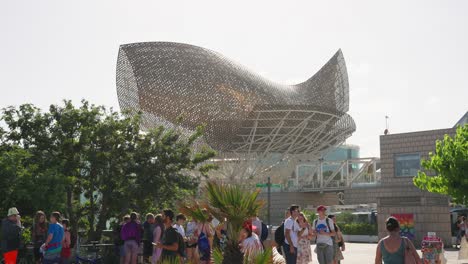A-group-of-teenagers-under-Frank-Gehry's-PEIX-monument-at-the-Port-Olimpic-in-Barcelona,-Spain