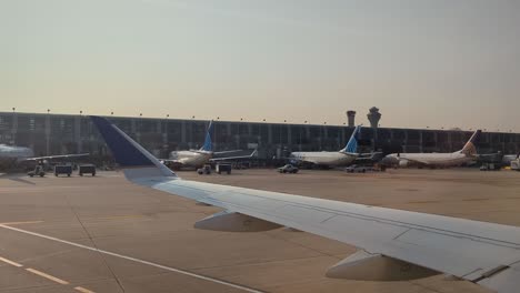 Vehículos-Terrestres-Del-Aeropuerto-Pasando-Por-Aviones-Estacionados-En-El-Aeropuerto,-Con-Vista-Parcial-Del-Ala-Del-Avión.