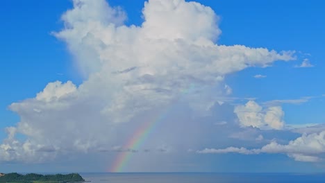 Ein-Wunderschöner-Regenbogen-über-Einer-Ruhigen-Blauen-Meereslandschaft-Mit-Einer-Großen,-Flauschigen-Weißen-Wolke,-Die-Darüber-Thront