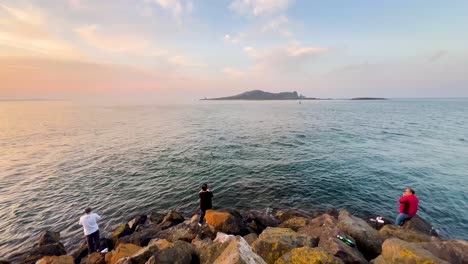 Fishing-at-Howth-Pier:-Serene-Sunset-Scene-with-Fisher,-Rocky-Coast,-and-Calm-Waters-by-the-Sea
