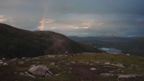 Female-Traveler-And-Pet-Dog-Hiking-On-Grytetippen-In-Norway-With-View-Of-Rainbow-In-Sunset-Sky