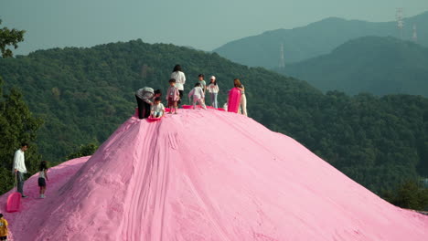 Kinder-Beim-Sandboarden,-Die-Nacheinander-Den-Rosafarbenen-Sandhügel-Auf-Der-Kräuterinsel-Mit-Malerischen-Bergen-Im-Hintergrund-Hinunterrutschen-–-Reiseziele-In-Südkorea