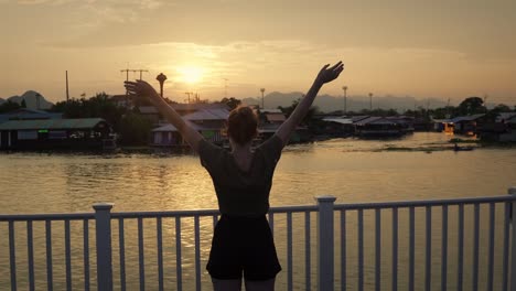 Young-Attractive-Female-Tourist-Standing-on-Harbor-Whard-with-Raised-Hands-at-Sunset-in-Thailand