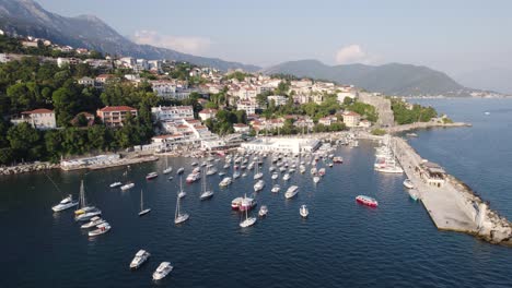 aerial:-Herceg-Novi-marina,-Montenegro,-with-sunlit-boats-and-mountain-backdrop