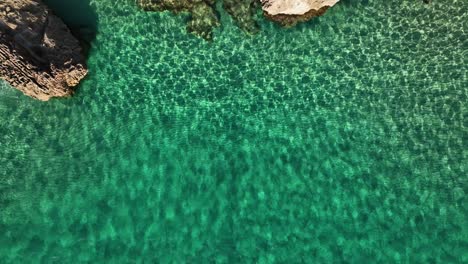 Top-down-drone-shot-of-shallow-crystal-clear-water-towards-a-rocky-coastline