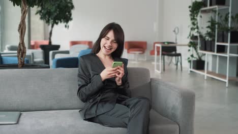 young-and-beautiful-woman-in-a-suit-sits-on-the-couch-in-the-business-center,-playing-a-mobile-game-on-her-smartphone-with-excitement,-smiling,-and-joyfully-celebrating-victory