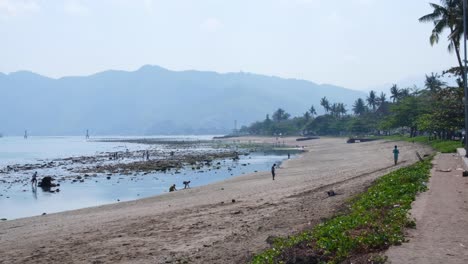 Grupos-De-Personas-En-La-Playa-De-Arena,-Haciendo-Ejercicio,-Jugando-Y-Buscando-Mariscos-En-Las-Aguas-Poco-Profundas-Del-Océano-En-La-Ciudad-Capital-De-Dili,-Timor-Oriental,-Sudeste-De-Asia