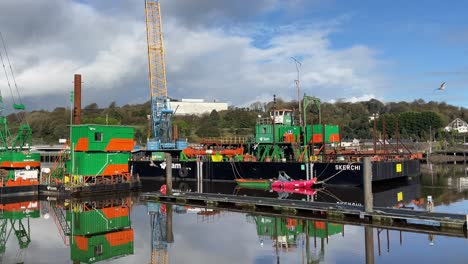 Construction-barge-on-a-calm-river-building-a-new-bridge-on-The-River-Suir-In-Waterford-Ireland-in-a-calm-clear-winter-day-with-birds-flying-past