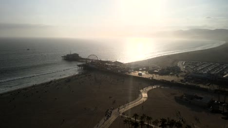 Una-Vista-Panorámica-Del-Parque-De-Diversiones-De-Santa-Mónica-Al-Atardecer,-Con-Las-Olas-Del-Mar-Rompiendo-En-La-Costa