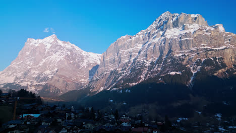 Aerial-reverse-dolly-pulls-away-from-snowy-grey-mountains-shading-the-village-of-Grindelwald-Switzerland
