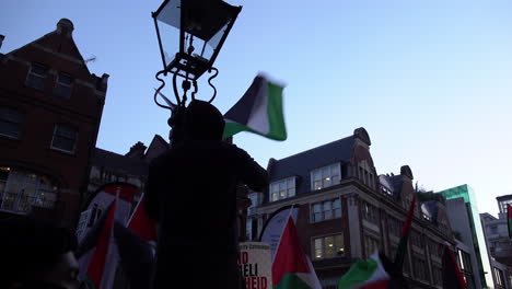 Ein-Mann-Mit-Schwarzem-Kapuzenoberteil-Schwenkt-Eine-Palästinensische-Flagge,-Nachdem-Er-In-Der-Abenddämmerung-Einen-Verzierten-Laternenpfahl-Vor-Der-Israelischen-Botschaft-Erklommen-Hat