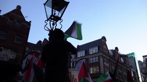 Un-Hombre-Vestido-Con-Una-Capucha-Negra-Ondea-Una-Bandera-Palestina-Después-De-Haber-Trepado-A-Una-Farola-Ornamentada-Frente-A-La-Embajada-De-Israel-Al-Atardecer.