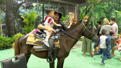 A-little-boy-sitting-on-a-horse-as-the-horse-handler-is-holding-him,-his-father-is-preparing-to-take-his-photo,-with-other-families-are-in-the-background-inside-an-amusement-park-in-Bangkok,-Thailand