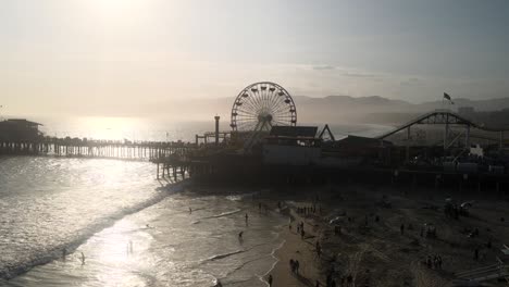Drone-Aerial-Cinematic-Shot-of-a-scenic-view-of-the-Santa-Monica-amusement-park-at-sunset,-with-ocean-waves-lapping-at-the-shoreline