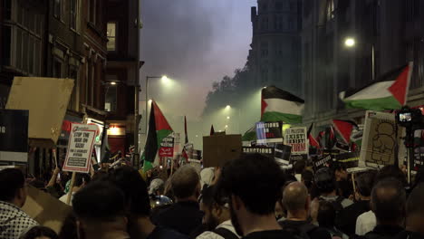 A-packed-crowd-of-people-hold-up-placards-and-flags-as-smoke-rises-up-from-colour-distress-flares-during-a-pro-Palestinian-protest-outside-the-Israeli-embassy-at-dusk