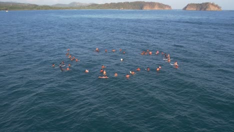 Drone-view-of-dozens-of-people-floating-in-the-sea-at-Samara-beach-carrying-surfboards-in-a-circle-formation,-Costa-Rica