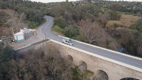 Aerial-tracking-shot-of-a-van-crossing-a-stone-bridge-in-Malaga,-Spain,-amid-lush-surroundings