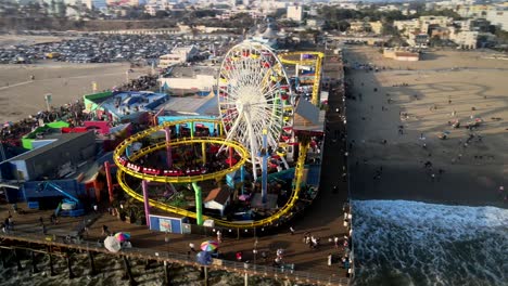 An-elevated-perspective-of-Pacific-Park-Pier-featuring-individuals,-a-roller-coaster-in-motion,-and-ocean-waves-breaking-on-the-shoreline