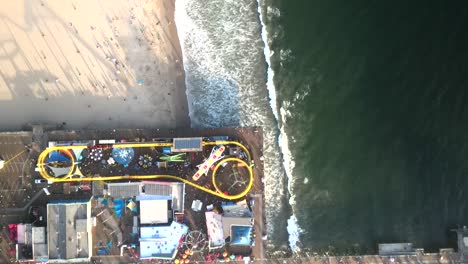 An-aerial-perspective-looking-down-upon-Santa-Monica's-Pacific-Park-Pier,-where-ocean-waves-gracefully-roll-onto-the-shoreline