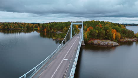 Dynamic-aerial-shot-of-a-suspension-bridge-crossing-a-lake-surrounded-by-an-autumn-forest-with-red,-green,-yellow-and-brown-trees-in-Valkeakoski,-Finland