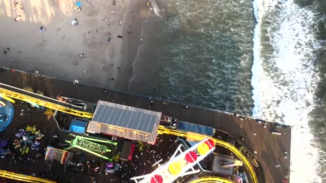 An-aerial-perspective-looking-down-upon-Santa-Monica's-Pacific-Park-Pier,-where-ocean-waves-gracefully-roll-onto-the-shoreline