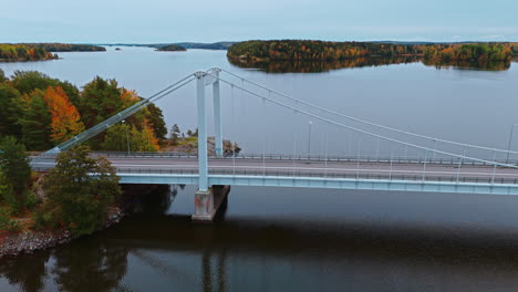 Orbiting-aerial-shot-of-a-suspension-bridge-crossing-a-lake-and-islands-surrounded-by-an-autumn-forest-with-green,-red-yellow-and-brown-trees