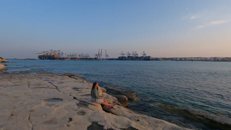 A-lady-sits-on-the-rocks-on-the-Mediterranean-coast,-with-the-port-of-Valletta-in-the-background