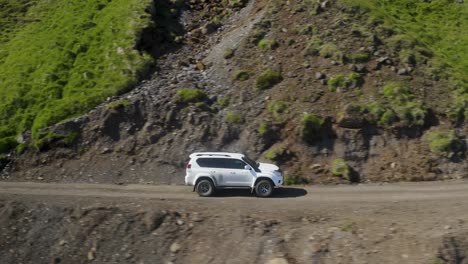 Aerial-drone-shot-of-white-suv-driving-on-volcanic-road-along-mountains-during-sunny-day-in-Iceland---Establishing-drone-shot