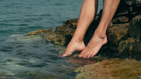 Man-dipping-toes-and-bare-feet-in-sea-water-at-rocky-shore