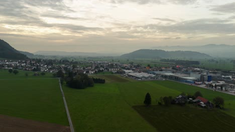 Aerial-backwards-shot-of-Bilten-Village-with-green-fields-during-cloudy-sunrise-in-Switzerland---Silhouette-of-mountains-in-background