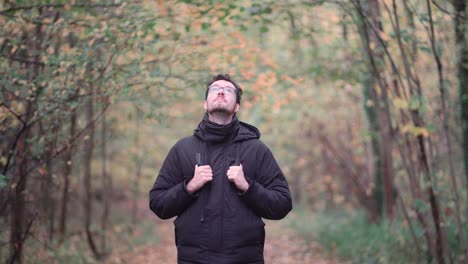Bearded-young-European-man-with-stylish-glasses-standing-amid-dense-forest,-intently-observing-the-mesmerizing-beauty-of-yellow-autumn-leaves-and-embracing-the-tranquility-of-nature
