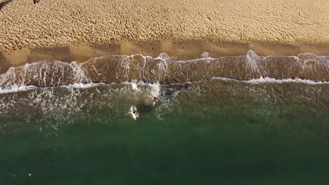 Zwei-Männer-Rennen-Ins-Wasser-Und-Schwimmen-Aus-Dem-Bild,-über-Ihnen-Dröhnt-Eine-Drohne