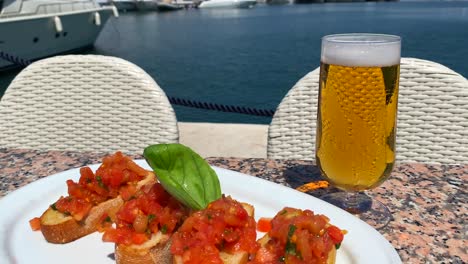 Classic-traditional-Italian-bruschetta-bread-with-tomatoes-and-olive-oil,-cold-beer-glass-and-view-on-luxury-yachts-in-Puerto-Banus-port-during-summer-in-Marbella-Spain,-tasty-food,-4K-shot