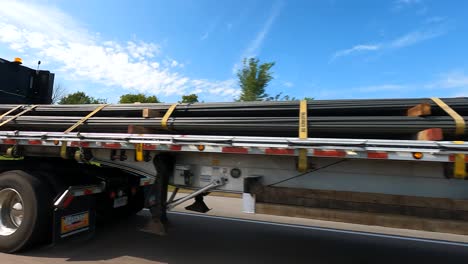 View-from-car-passing-flatbed-truck-transporting-steel-pipes-on-sunny-day