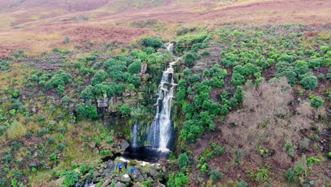 Majestic-Yorkshire-moor-cascade,-aerial-view:-water-flows-over-boulders,-dropping-into-a-deep-blue-pool,-with-hikers-standing