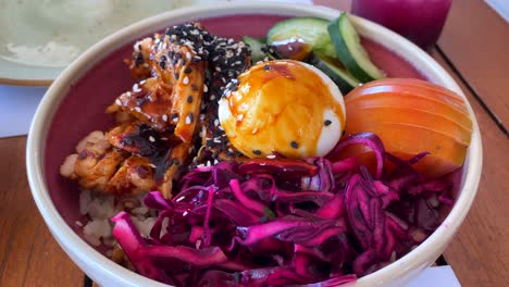 Delicious-asian-bowl-with-chicken,-rice,-egg,-purple-cabbage,-tomatoes,-cucumbers-and-black-sesame-seeds-at-a-restaurant,-4K-shot