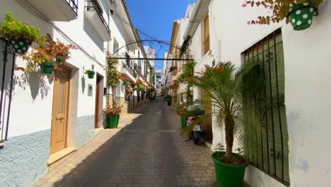 Walking-through-a-little-scenic-street-in-Estepona-old-town-with-houses,-colorful-flower-pots-and-beautiful-balconies,-sunny-day-in-Andalusia-Spain,-4K-shot