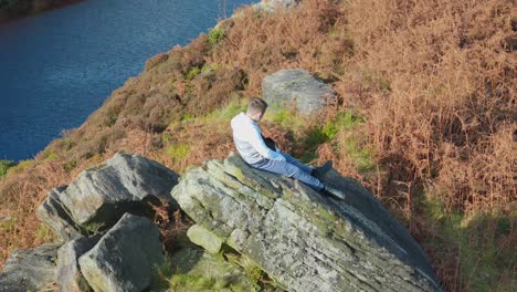 Aerial-video-gently-orbits-a-pensive-young-boy-perched-on-a-massive-rock-outcrop-in-the-moorland-setting