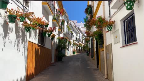 Walking-through-scenic-Estepona-old-town-in-a-narrow-Spanish-street-with-houses,-colorful-flower-pots-and-beautiful-balconies,-sunny-day-in-Andalusia-Spain,-4K-shot