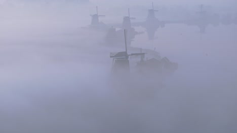 Row-of-traditional-windmills-at-the-Netherlands-during-foggy-sunrise,-aerial