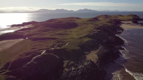 Aerial-view-circling-Ynys-Llanddwyn-Welsh-island-with-shimmering-ocean-and-misty-Snowdonia-mountain-range-across-the-sunrise-skyline