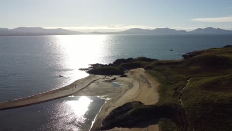 Aerial-view-rising-above-Welsh-Ynys-Llanddwyn-island-beach-with-shimmering-ocean-and-misty-Snowdonia-mountain-range-across-the-sunrise-skyline