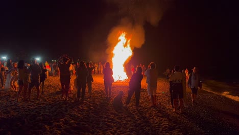 Gente-Bailando-En-El-Tradicional-Festival-De-Verano-De-Hogueras-En-La-Playa-En-La-Celebración-De-San-Juan-En-Marbella-España,-Disfrutando-De-Una-Fiesta-Divertida,-Un-Gran-Fuego-Ardiente-Y-Llamas-Calientes-Por-La-Noche,-Tomas-De-4k