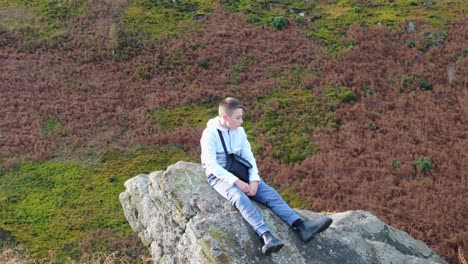 Aerial-footage-revolves-around-a-thoughtful-young-boy-seated-on-a-substantial-rock-outcrop-in-the-midst-of-the-moorland