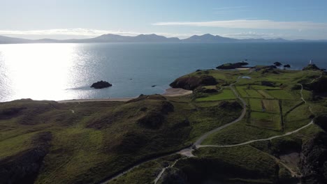 Aerial-view-above-Ynys-Llanddwyn-Welsh-island-with-shimmering-ocean-and-misty-Snowdonia-mountain-range-across-the-sunrise-skyline